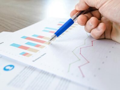 A close-up of a hand with a pen analyzing data on colorful bar and line charts on paper.